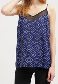 Svart linne med ett blått blommönster i diamantform, med en genomskinlig V-formad halsringning och tunna justerbara axelband. Slät tygtextur.