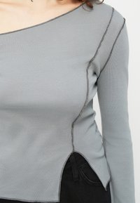 Γυναίκα που φοράει ένα ανοιχτό γκρι ασύμμετρο μακρυμάνικο τοπ με εκτεθειμένες ραφές και πλευρική σχισμή, συνδυασμένο με μαύρο παντελόνι.