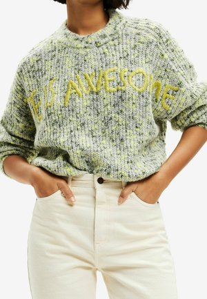 Osoba nosící zelený a šedý pletený svetr se žlutě vyšitým nápisem "BE IS AWESOME", doplněný kalhotami s vysokým pasem v béžové barvě.