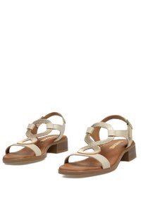 Sandalias de tacón beige con punta abierta, tira en el tobillo y detalles decorativos trenzados, con una plantilla marrón texturizada y tacón bajo.