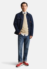 Homme debout portant une veste en jean foncée, un sweat beige, une chemise blanche, un jean bleu et des baskets rouges sur un fond blanc uni.