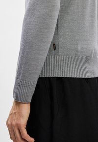 Sivkasta pletena jopica z rebrastim robom in manšetami, z teksturiranim telesom in majhnim blagovnim oznako na strani.