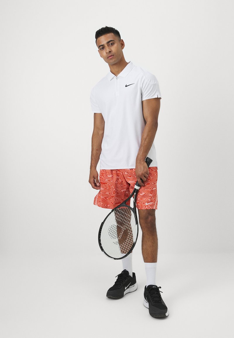 Biała koszulka polo z czarnym logo, pomarańczowe spodenki w wzory, czarne sneakersy i białe skarpetki, trzymająca rakietę tenisową.