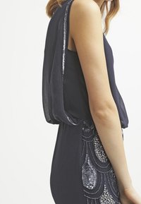 Robe marine avec des manches en tissu transparent, ornée de motifs de perles scintillants le long de l'ourlet. Texture douce et design fluide.