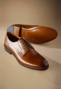 Zapatos de vestir de cuero marrón con un acabado suave, diseño con cordones, puntera redondeada y un detalle de suela de goma negra contrastante.