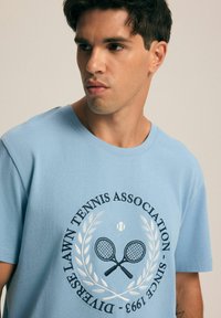 Jasnoniebieski bawełniany t-shirt z okrągłym dekoltem, krótkimi rękawami oraz grafiką przedstawiającą skrzyżowane rakiety tenisowe i wieniec laurowy.