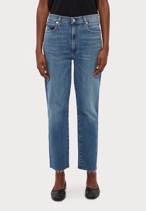 Person trägt blaue Jeans mit mittlerer Taille und geradem Bein, mit rohen Säumen und schwarzen flachen Schuhen, steht vor einem einfarbigen Hintergrund.