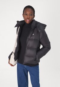 Black puffer jacket with a hood featuring a logo, closed zipper, and quilted texture. Paired with a black turtleneck and blue jeans.