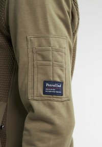 Olijfgroene jas met gestructureerd schouderpaneel, voorzien van een vierkante zak op de mouw en een blauw label met rode accenten.