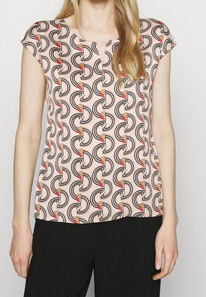 Mujer que lleva una blusa beige de manga corta con un patrón geométrico repetido en negro, naranja y rojo, combinada con pantalones negros.