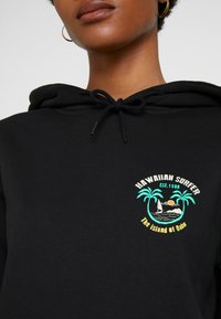 Juodas gobtuvas su siuvinėta palmėmis, jachta ir saule, ant kurios yra užrašas "Hawaiian Surfer EST. 1986" ryškiomis spalvomis.