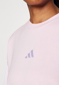 Svetlo roza majica s dolgimi rokavi in krožnim izrezom, mehka tekstura, z majhnim lila Adidas logotipom na levi strani prsnega dela. Preprost, vsakdanji dizajn.