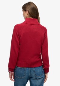 Rood gebreide trui met een hoge kraag, geribbelde manchetten en zoom, gedetailleerde naden en een losse pasvorm, gecombineerd met blauwe spijkerbroek.