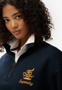 Marineblauwe rits-sweater met een geel geborduurd logo en kroonontwerp, met een hoge kraag over een wit shirt met zwarte accenten.