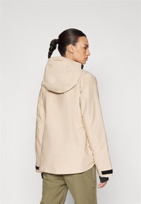 Beige vanntett jakke med hette, svarte mansjetter og avslappet passform. Har en logo ved halsen og en glatt tekstur.