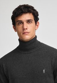 Maglione a collo alto di color grigio scuro realizzato in tessuto a maglia, con un design aderente e un piccolo logo sul lato sinistro del petto. Texture liscia.