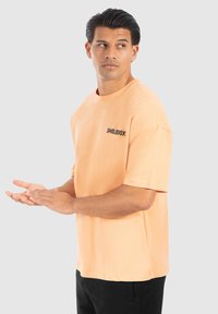 Kortærmet, løst siddende t-shirt i blød ferskenfarvet stof, med et sort "SMILODOX" logo på venstre brystområde.