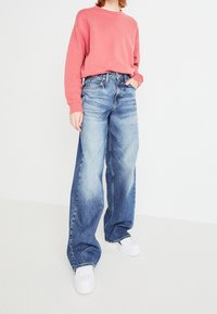 Osoba v bledě modrých širokých džínech, korálovém svetru a bílých teniskách, stojící s jednou rukou v kapse na jednobarevném pozadí.