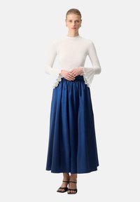 White long-sleeve top with ruffled bell sleeves paired with a floor-length, dark blue, gathered waist skirt, complemented by black strappy sandals.