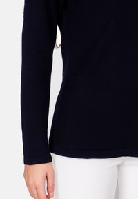 Pull-over en maille bleu marine à manches longues avec une texture douce, doté d'un design ajusté et d'un ourlet légèrement évasé. Main visible, portant un pantalon blanc.