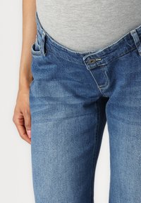 Denim kavbojke v srednjem modrem wash-u. Značilnosti vključujejo gumbasto zapiranje, pet žepov in rahlo zbledelost na stegnih. Tkanina iz mešanice bombaža.