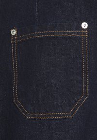 Bolsillo de unos vaqueros de mezclilla con forma rectangular, color azul oscuro, costuras naranjas en contraste y detalle de botón metálico cerca de la esquina superior.