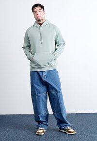 Helles grünes Hoodie mit einer Fronttasche und einem halben Reißverschluss, kombiniert mit lockeren blauen Jeans. Das Bild zeigt weiße und braune Sneakers.
