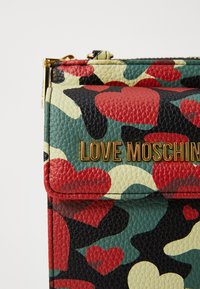 Textured faux leather handbag features a camouflage pattern with red and green hearts. Gold "LOVE MOSCHINO" logo accents the front.