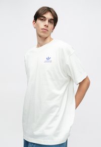 Balta kokvilnas t-krekls ar brīvu piegriezumu, uz krūtīm ir neliels purpura Adidas logo un īsās piedurknes.