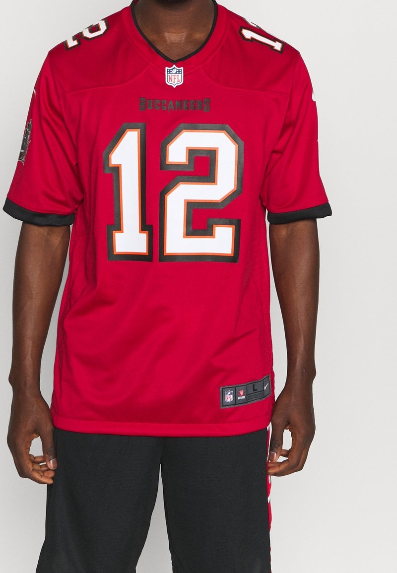 Homme portant un maillot rouge des Tampa Bay Buccaneers NFL avec le numéro 12, associé à un short noir, debout sur un fond blanc uni.