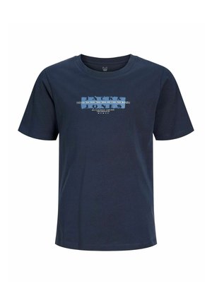 Tienerjongen staat, draagt een marineblauw grafisch t-shirt, olijfgroene cargobroek en witte sneakers, kijkt opzij tegen een effen achtergrond.