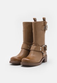 Ljusbruna läderstövlar till mitten av vaden med remmar, silverfärgade spännen, rundade tår och låga klackar mot en vit bakgrund.