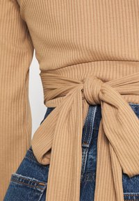 Top beige canelado com um nó na cintura, combinado com jeans azul denim com detalhes desgastados e costura amarela.