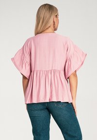 Różowa bluzka peplum z krótkimi, falbaniastymi rękawami, z marszczoną talią i luźnym krojem. Tkanina wydaje się lekka i lekko teksturowana.