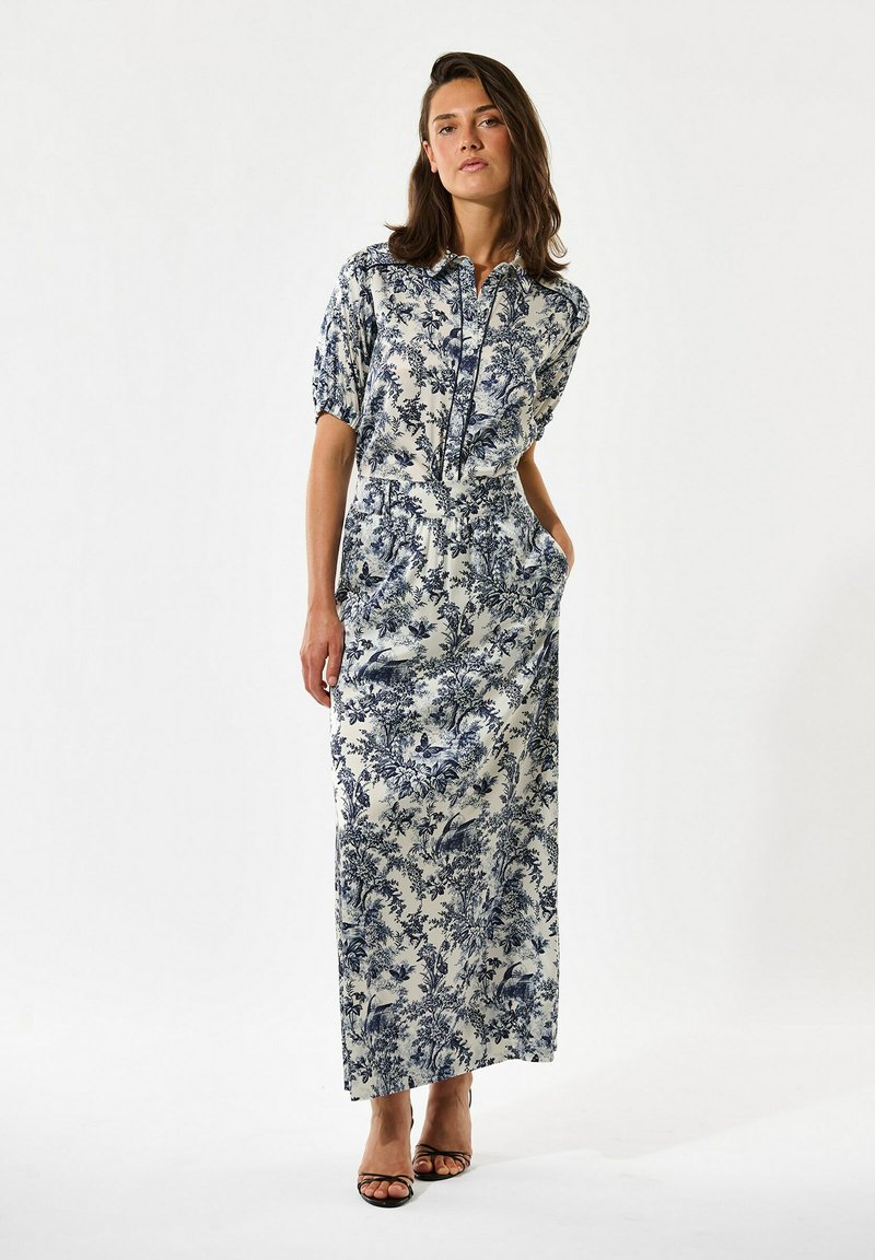 Vrouw staand in een lange, korte mouw witte jurk met donkerblauwe bloemen- en vogelprint, zwarte sandalen met bandjes, handen in de zakken.