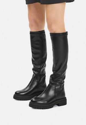 Botas negras de cuero hasta la rodilla con suelas gruesas de goma, usadas con piernas desnudas sobre un fondo blanco liso.