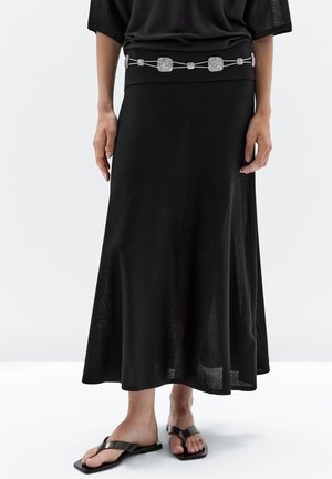 Une personne porte une tenue noire ample avec une ceinture décorative. Elle se tient pieds nus dans un décor minimaliste et gris clair.