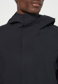 Czarna wodoodporna kurtka z wysokim kołnierzem i kapturem. Charakteryzuje się detalami szwów oraz matowym wykończeniem. Gładka faktura i minimalistyczny design.