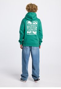 Sweat à capuche vert avec un motif graphique blanc au dos, représentant des visages abstraits. Associé à un jean large bleu clair et des chaussures noires.