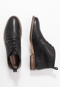 Botas de tobillo de cuero negro con acabado suave, punta redonda y diseño con cordones. Suela de goma marrón claro con detalles de costura y pestaña en el talón.