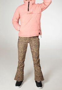 Personne portant une doudoune rose et un pantalon à imprimé léopard, debout avec une main dans la poche et l'autre levée.