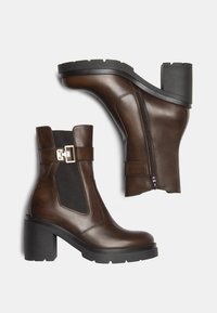 Des bottines en cuir marron présentent un talon noir épais, des panneaux latéraux élastiques et une boucle décorative sur la sangle. Texture lisse avec une fermeture éclair.