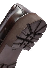Zapato de cuero marrón oscuro con punta redondeada y detalles de costura; cuenta con una suela de goma gruesa y texturizada para agarre y durabilidad.