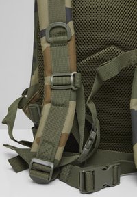 Kamufliažinis kuprinės dirželis su minkštu tinkleliu atloše, reguliuojamais sagtimis ir plastikiniais užsegimais pilkame fone.