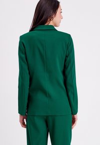 Green blazer with a tailored fit, sharp lapels, and long sleeves. Smooth fabric with a streamlined back and minimal detailing.