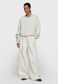 Sweat-shirt gris clair avec l'inscription "TODAYS" en lettres blanches, associé à un pantalon taille haute à jambes larges de couleur blanc cassé et des chaussures foncées.