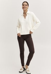 Hvid striktrøje med krave, forlommer og ribbet kant, parret med brune slim-fit bukser og hvide loafers med guld accenter.
