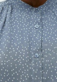 Blouse bleu clair à pois blancs, avec un col arrondi et trois boutons bleus le long de la patte de boutonnage avant. Tissu doux et lisse.