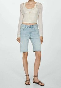 Lehký, průsvitný krémový kardigan přes bílý top, kombinovaný s světlemodrými džíny šortkami, které mají třepený lem a klasický pětizubý design.