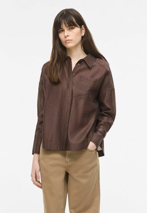 Jonge vrouw met lang bruin haar die een losse donkerbruine blouse met knopen en beige broek draagt, staand met één hand in haar zak.